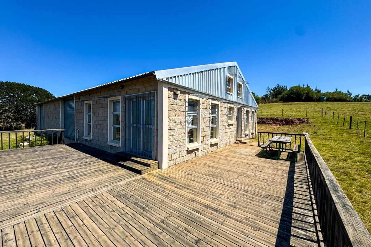 A rear view of a stone-clad farm building featuring an expansive timber deck, metal roofing, and security gates, set against a rural landscape.