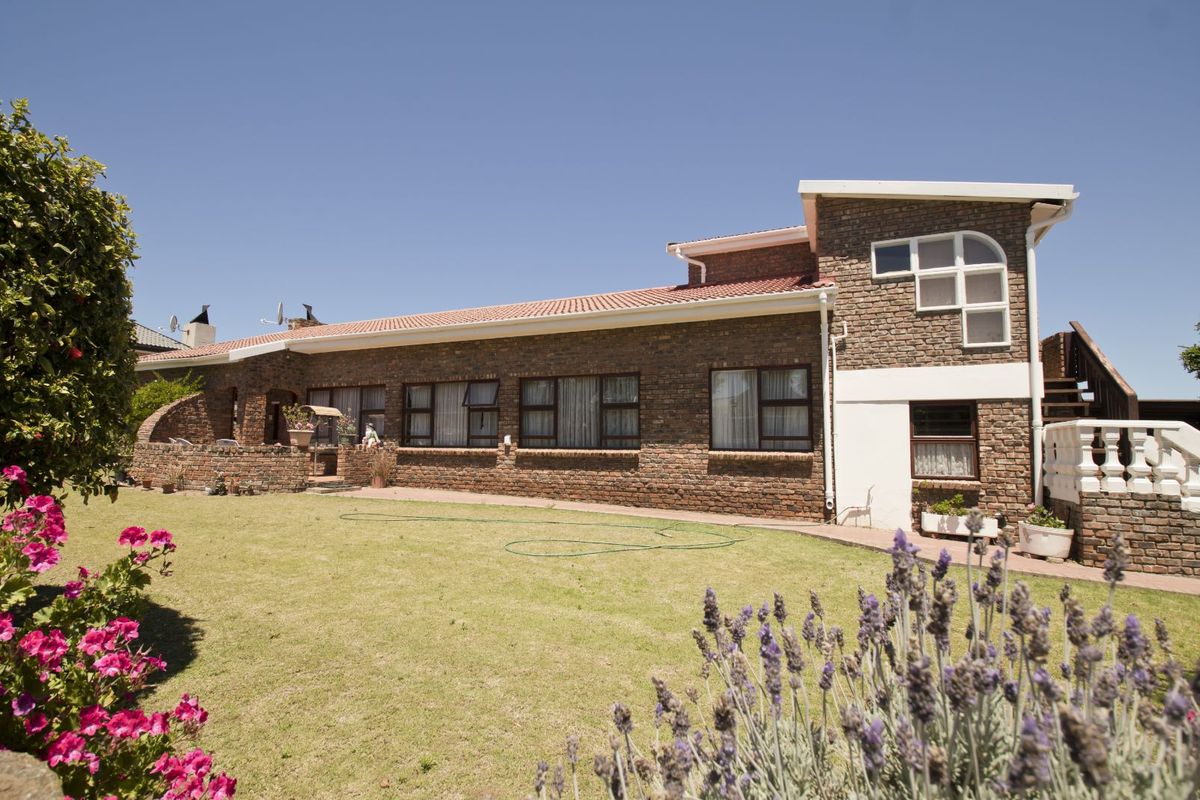 A view of the rear exterior of a brick house featuring a large lawn, tiled roof, and a raised patio area.