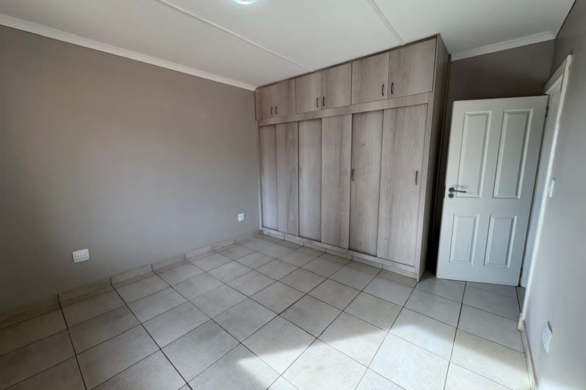 A clean, empty bedroom featuring neutral tiled flooring, built-in wooden cupboards with overhead storage, and a classic white panel door.
