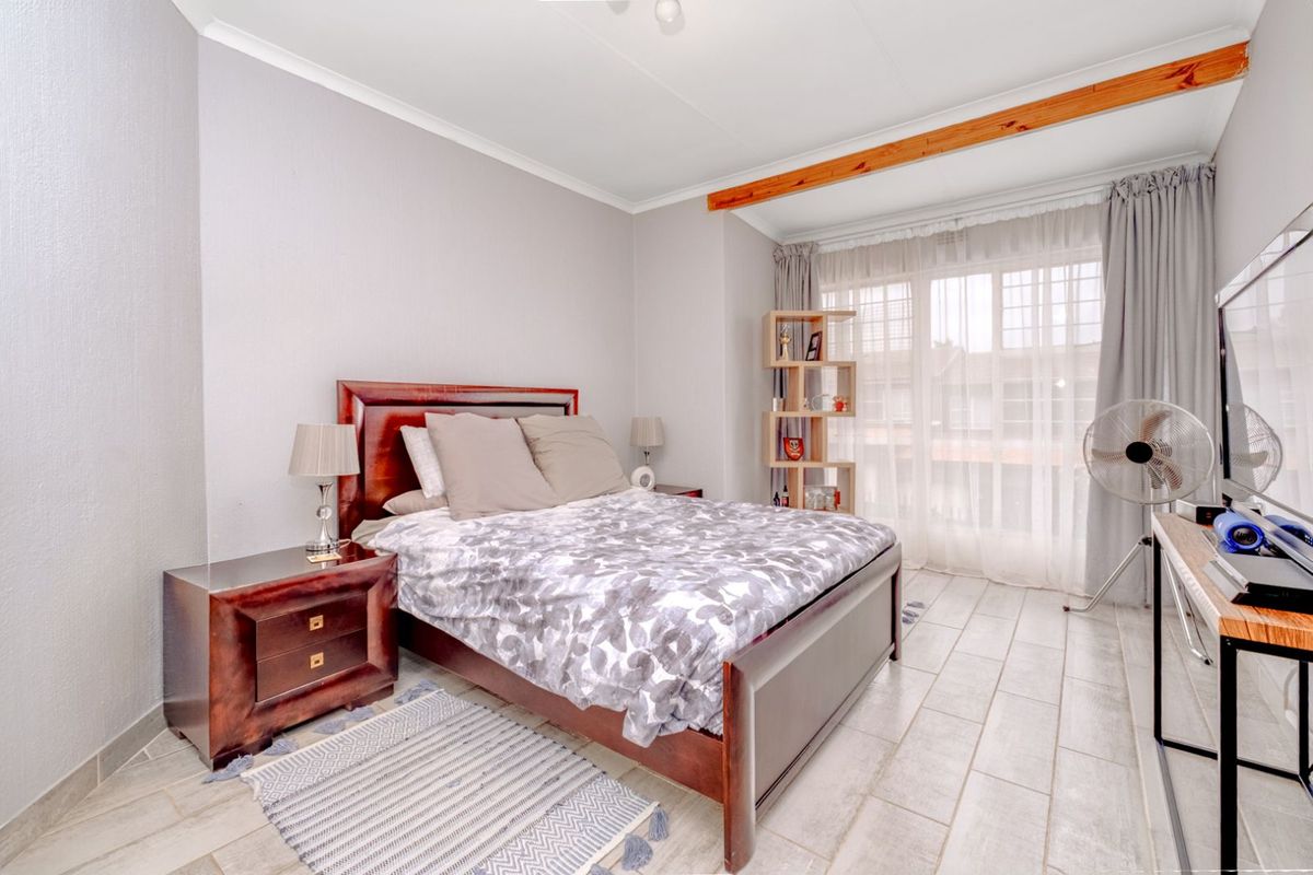 A well-lit bedroom featuring tiled flooring, a decorative wooden ceiling beam, and large windows with sheer curtains allowing for natural light.