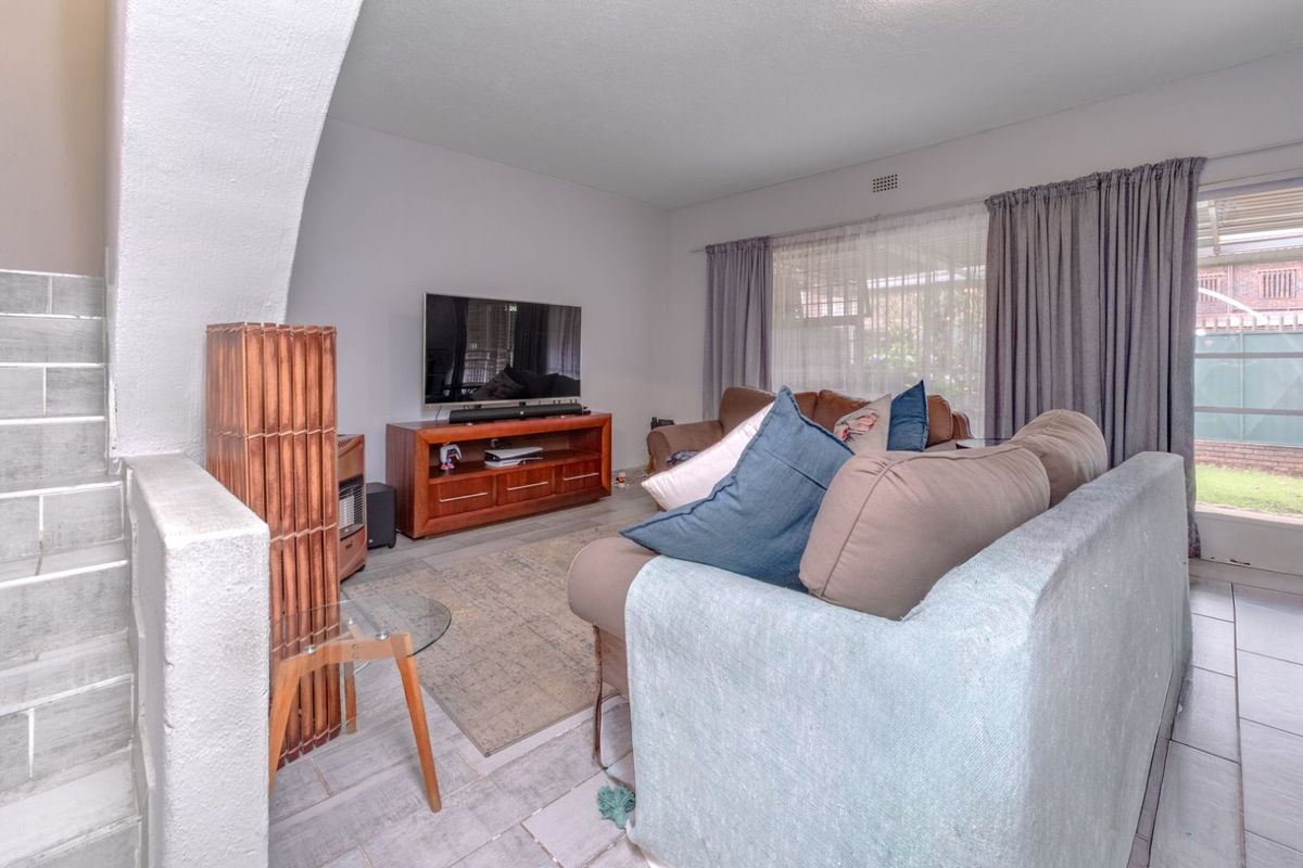 A living room featuring tiled flooring, an arched staircase partition, and large windows with curtains, providing access to the outdoor area.