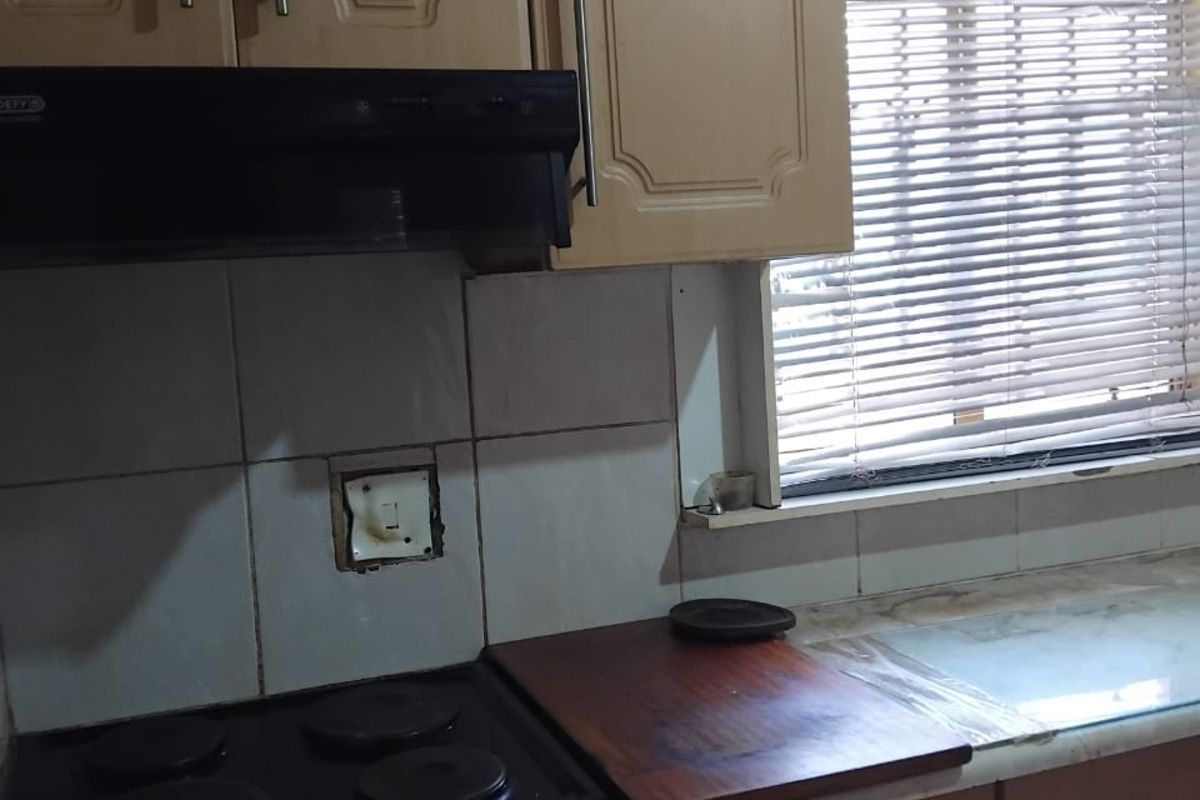 A dated kitchen area featuring built-in cabinetry, a four-plate electric hob, extractor fan, and a window with fitted blinds.