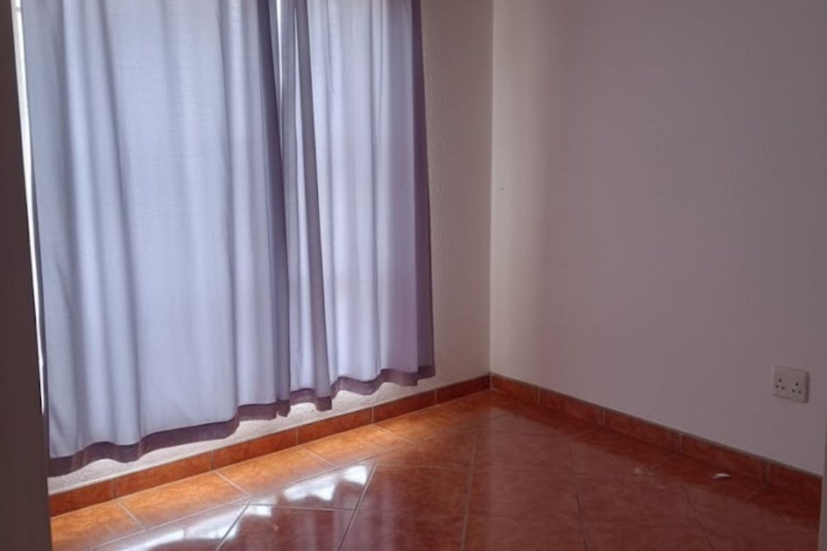 A simple, empty bedroom featuring terracotta-style floor tiles and a window with curtains.