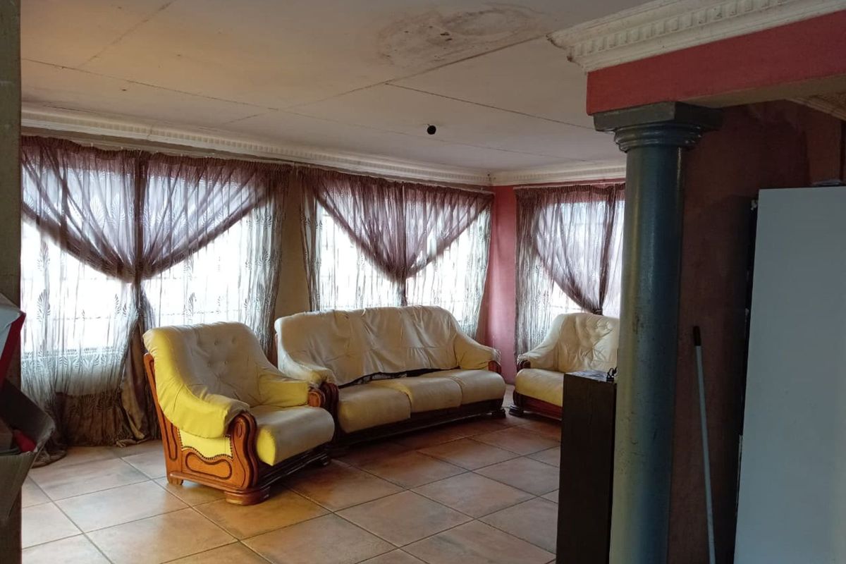 A living room area featuring tiled flooring, decorative columns, and large windows with curtains, showing signs of wear and ceiling water damage.