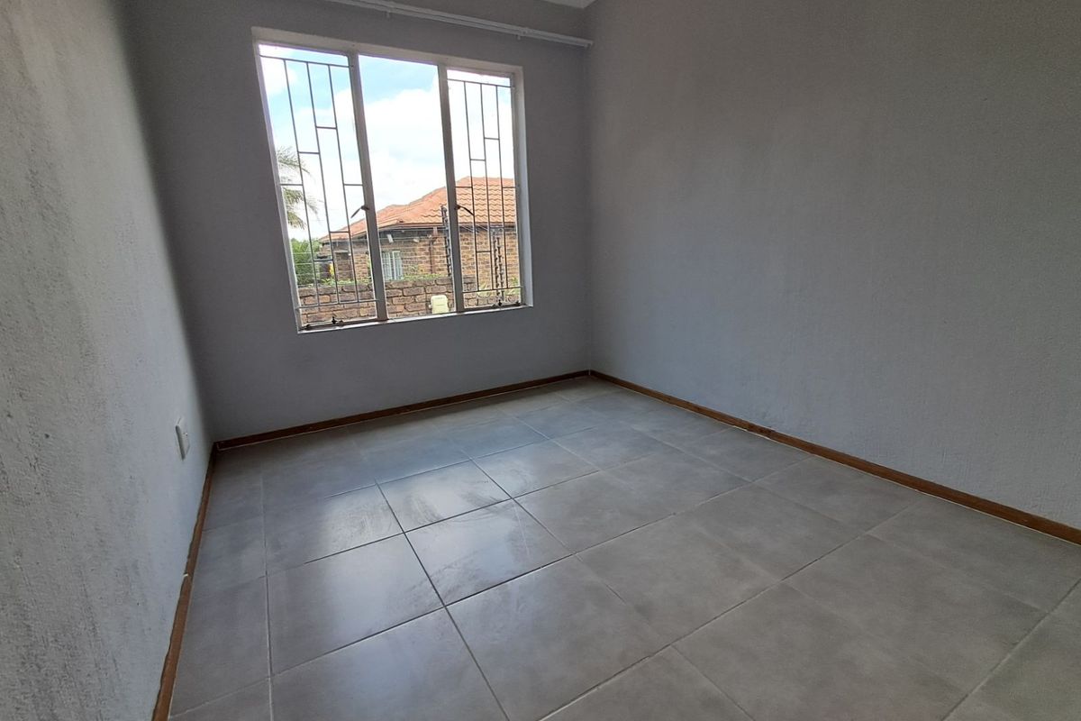 A vacant bedroom featuring neutral grey tiled flooring, plain walls, and a window fitted with security bars.