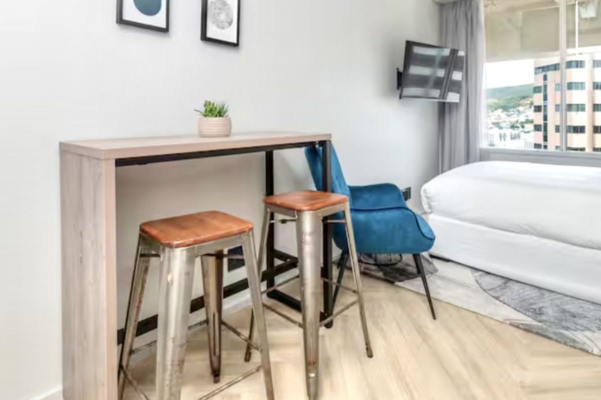 A modern bedroom featuring light wood-look flooring, large windows with city views, and a wall-mounted television.