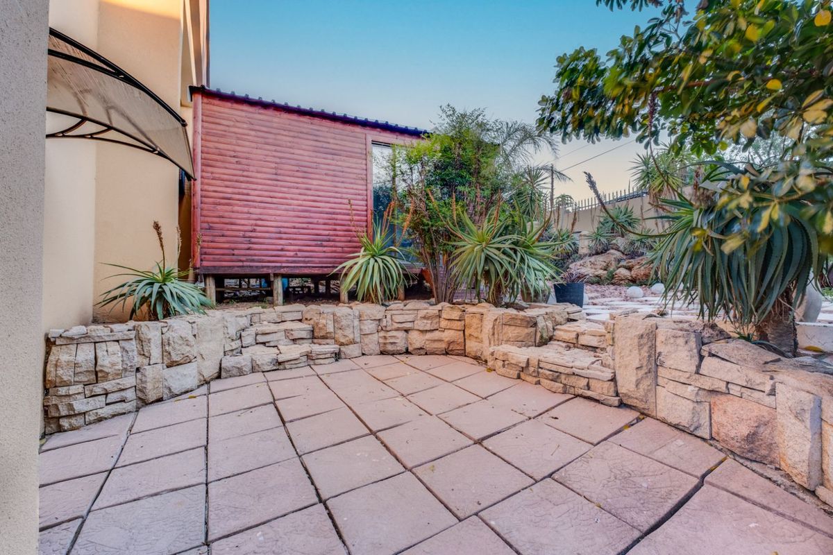 A rear garden area featuring a paved patio, stone retaining wall, and a raised wooden structure set against a landscaped backdrop.