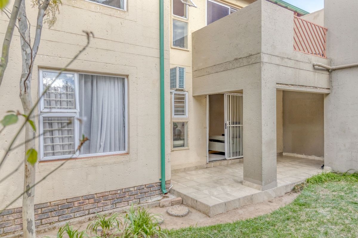 A view of the apartment's rear exterior, featuring a covered patio area, tiled flooring, and access to the interior through a security gate.