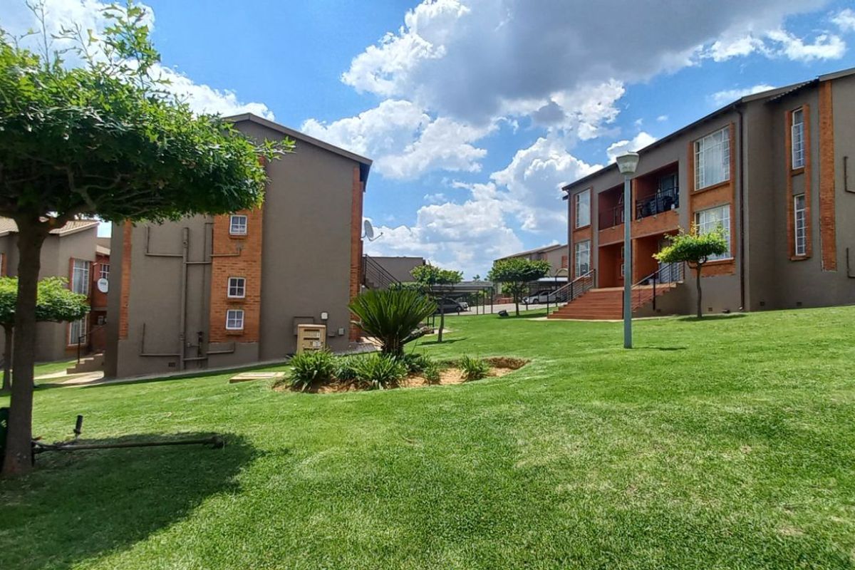 A view of the apartment complex grounds featuring well-maintained lawns, landscaped garden beds, and multi-story residential buildings with brick and render facades.