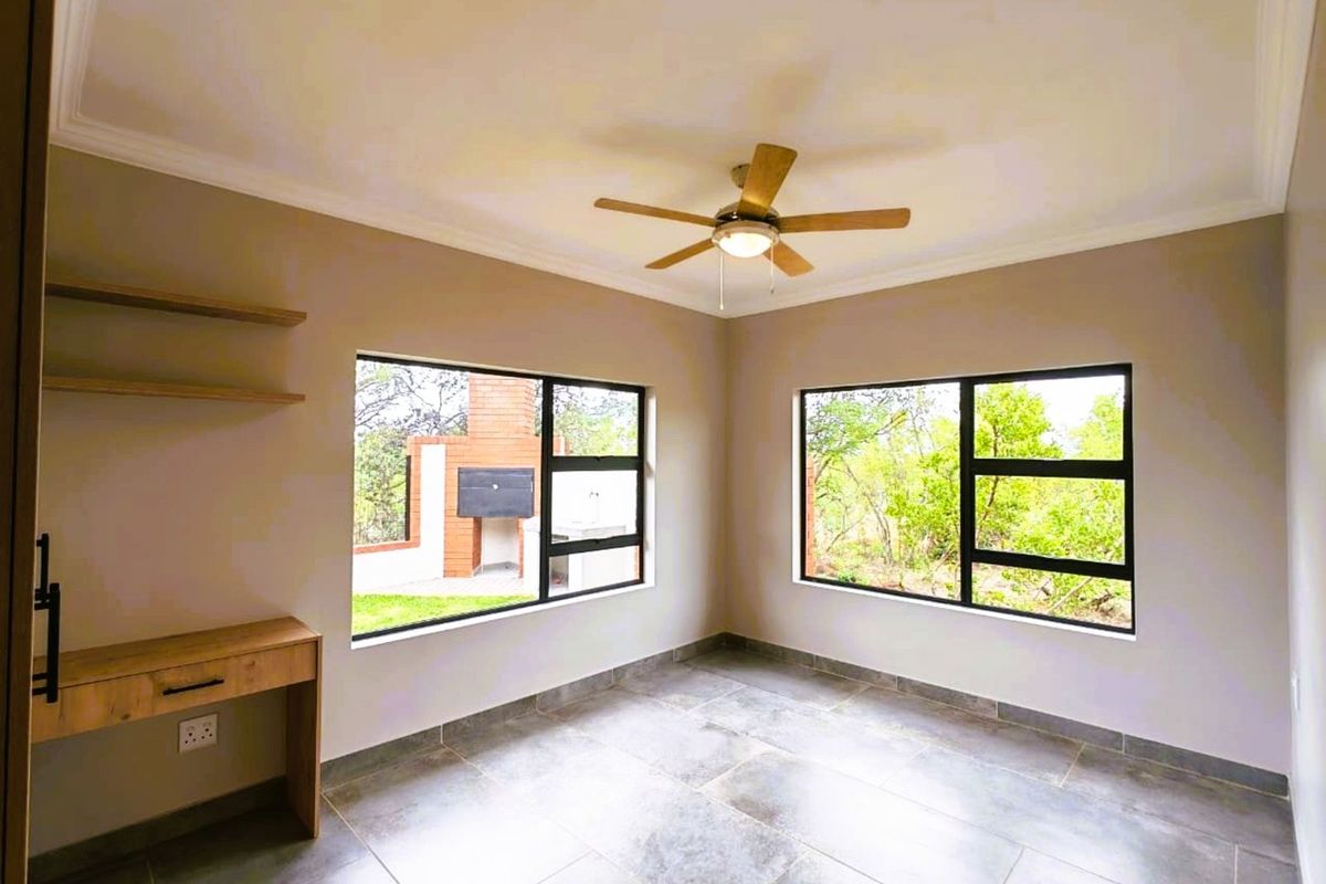 A bright bedroom featuring large windows with black frames, tiled flooring, a ceiling fan, and a built-in wooden desk with shelving.
