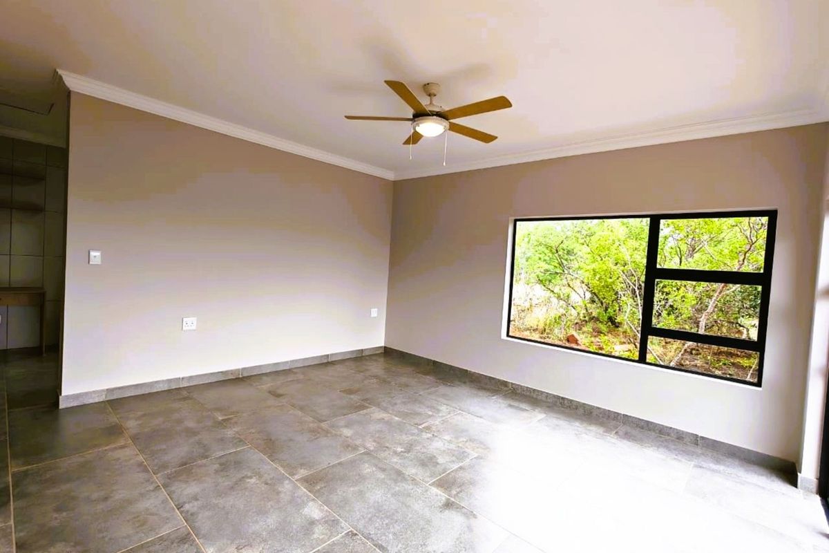 A bright, empty living room featuring large windows with garden views, modern tiled flooring, and a ceiling fan.