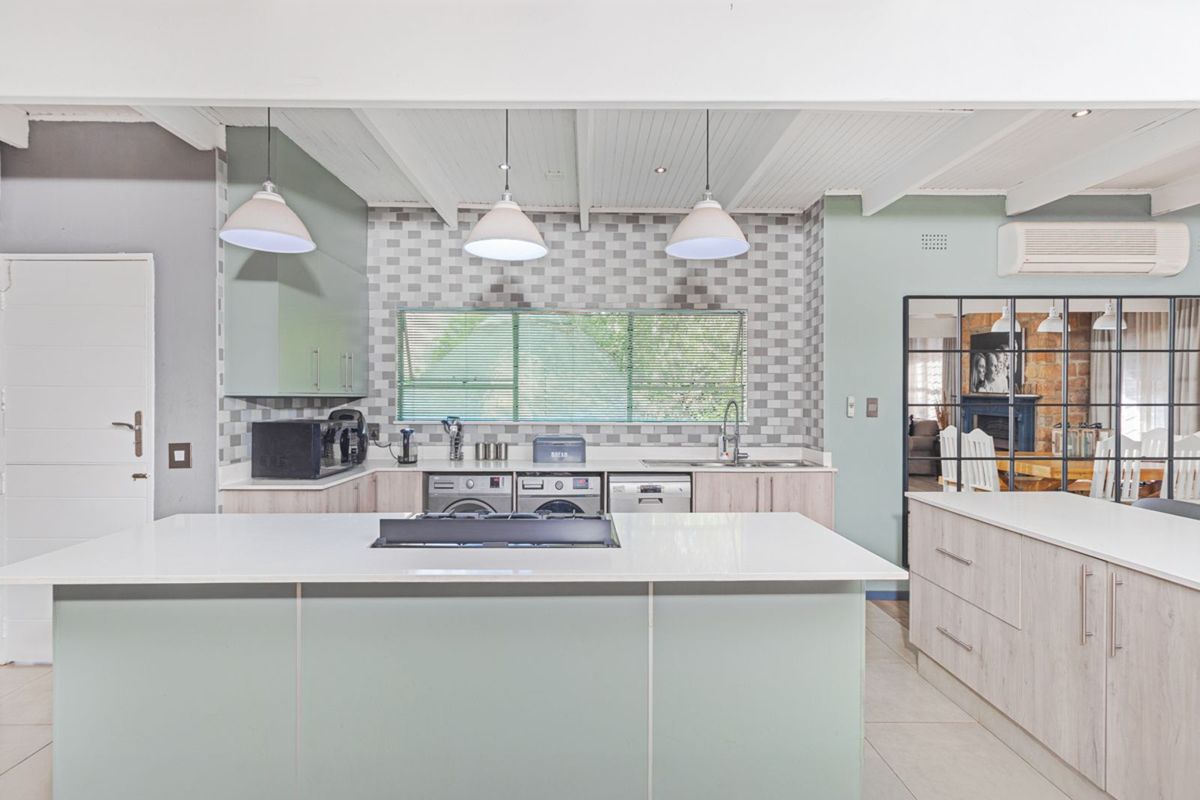 Modern kitchen featuring a central island with integrated cooktop, light-toned cabinetry, tiled backsplash, and exposed beam ceiling, seamlessly connecting to the adjacent living area.