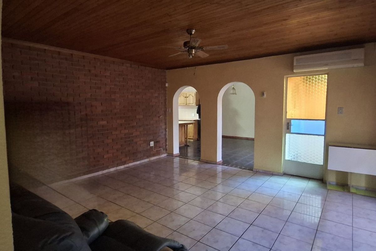 Spacious living area featuring a rustic exposed brick wall, tiled flooring, timber-clad ceiling, and arched doorways leading to the kitchen area.