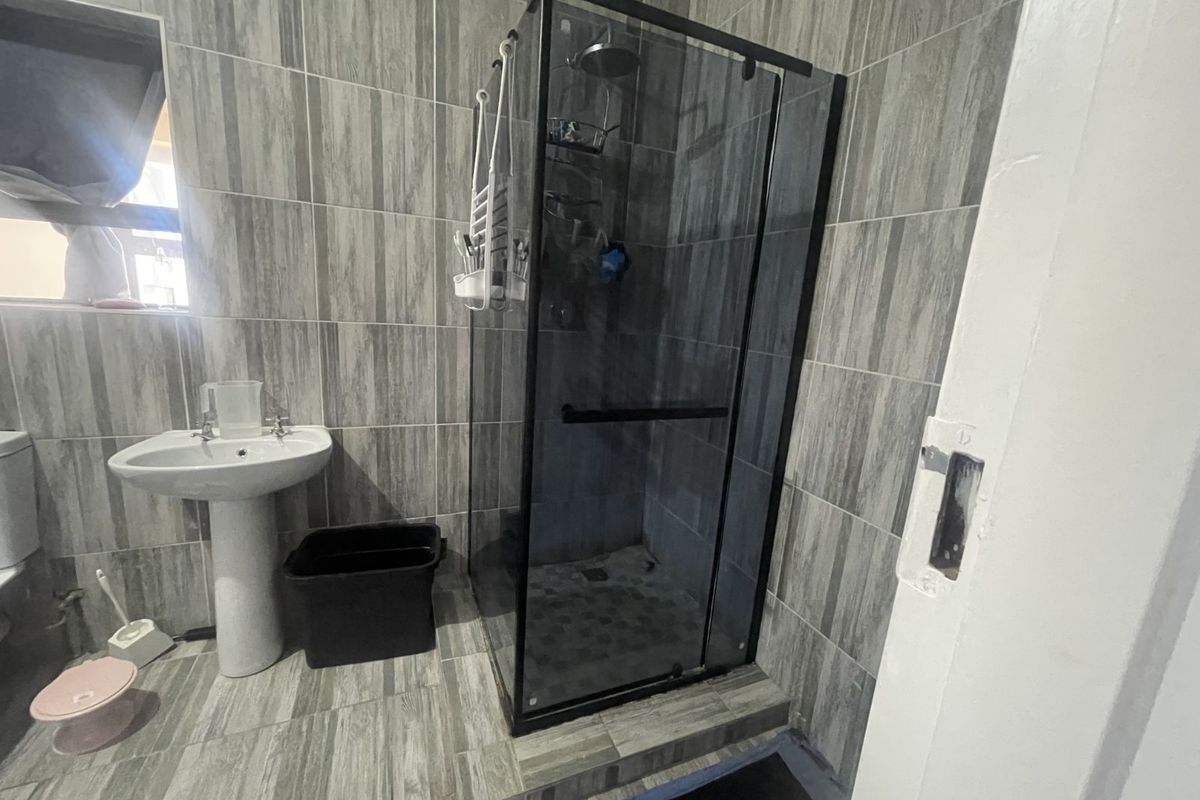 A modern bathroom featuring grey wood-look floor and wall tiles, a glass-enclosed shower with black frame, and a white pedestal basin.