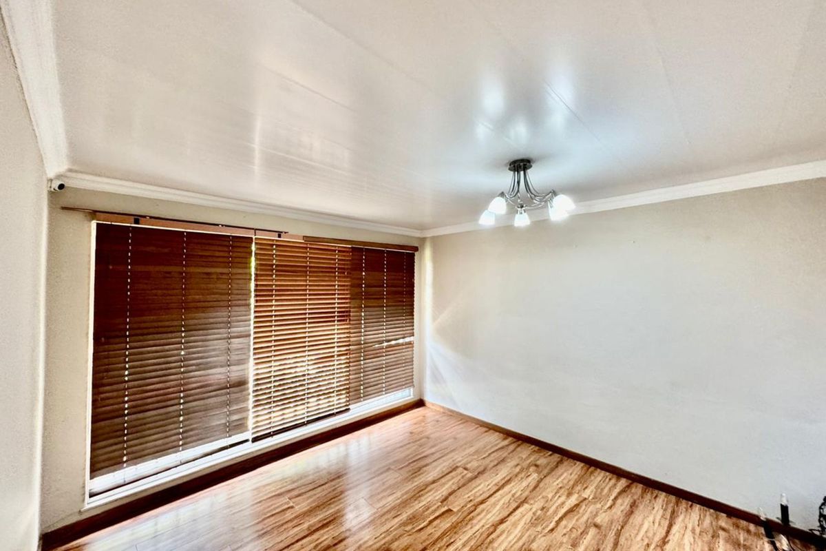 A bright, empty bedroom featuring wood-look laminate flooring, large windows with fitted wooden blinds, and a central ceiling light fixture.