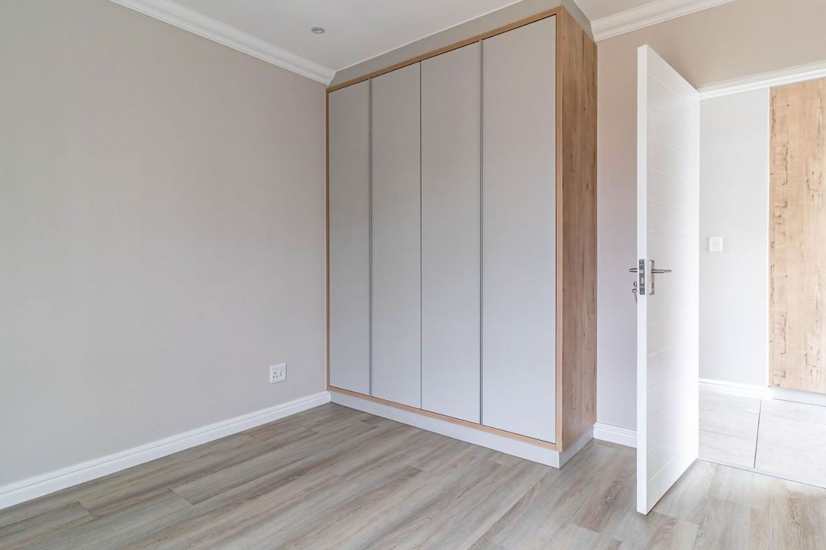 A bright bedroom featuring modern built-in wardrobes with light grey doors and wood-look trim, complemented by wood-look laminate flooring and crown molding.