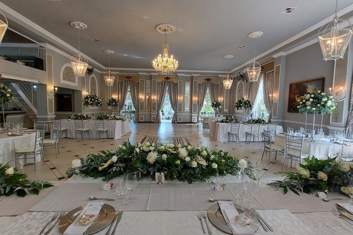 Spacious event hall featuring elegant crown molding, decorative ceiling medallions, classic chandeliers, and large windows with draped curtains, suitable for formal gatherings.