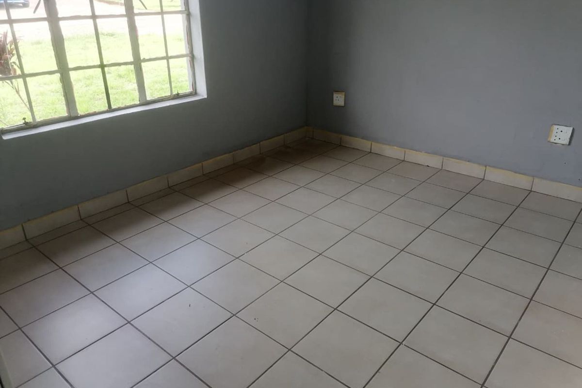 A vacant bedroom featuring neutral grey walls, tiled flooring, and a large window providing natural light and a view of the front garden.