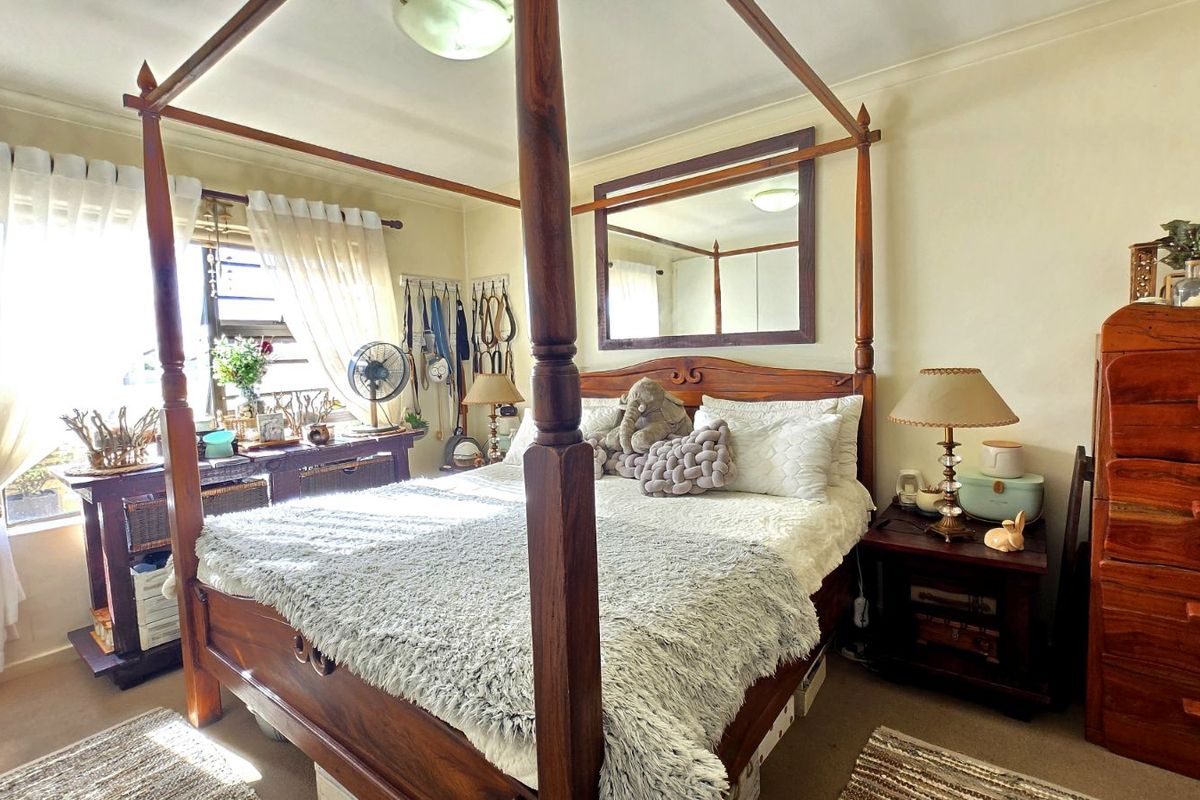 A well-lit bedroom featuring a four-poster bed frame, wall-mounted mirror, and large window with natural light.