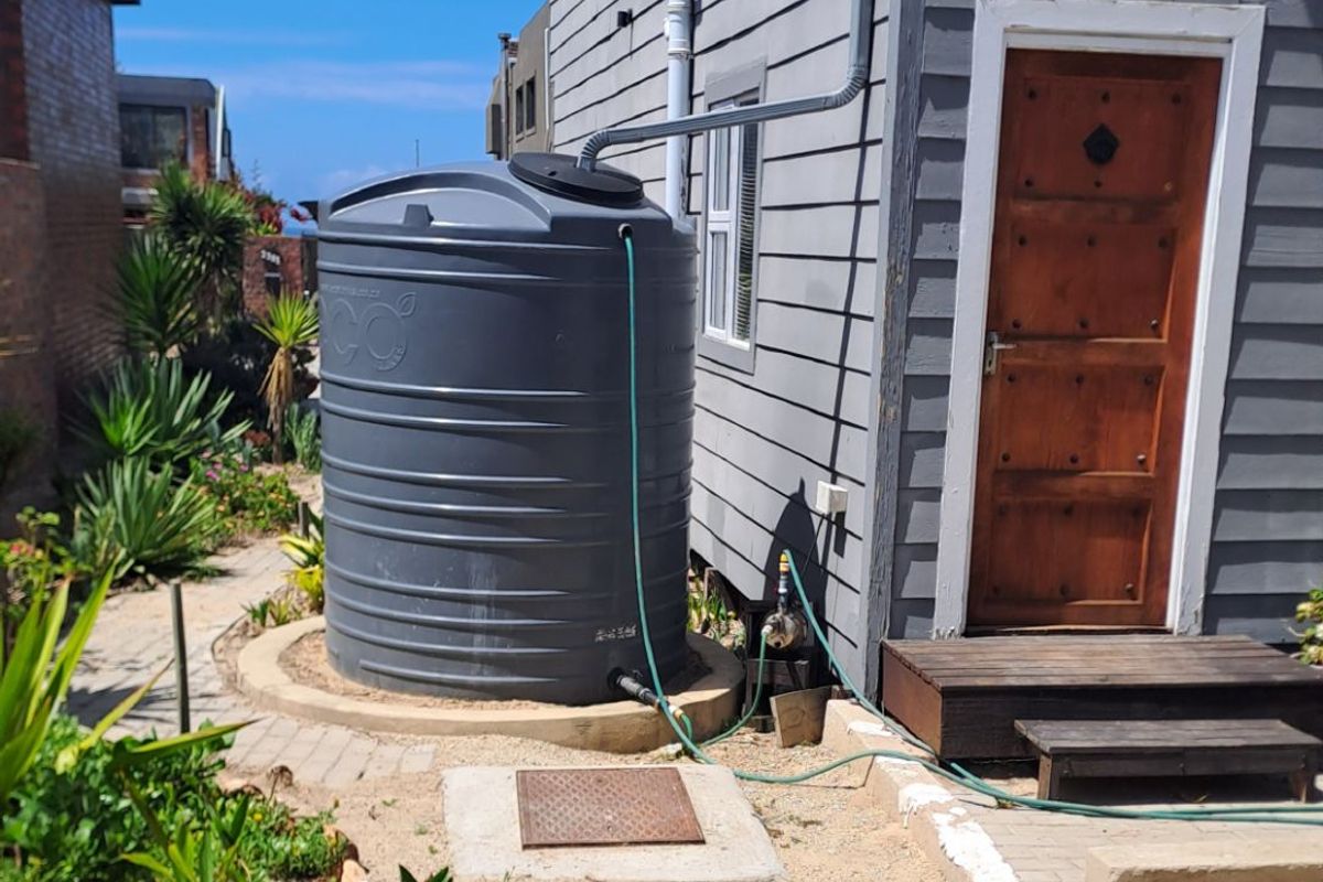 Exterior view of the property side featuring a large grey rainwater storage tank, downpipe system, and a timber access door with steps.