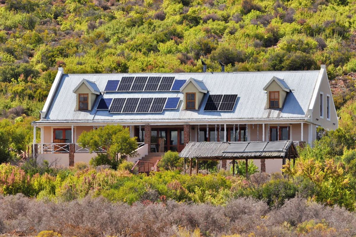 A detached house featuring a corrugated metal roof with integrated solar panels, dormer windows, and a covered outdoor patio area set in a natural landscape.