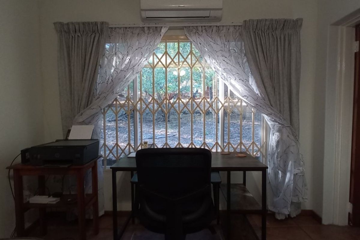 A functional bedroom or office space featuring a wall-mounted air conditioning unit and a window secured with metal security bars.
