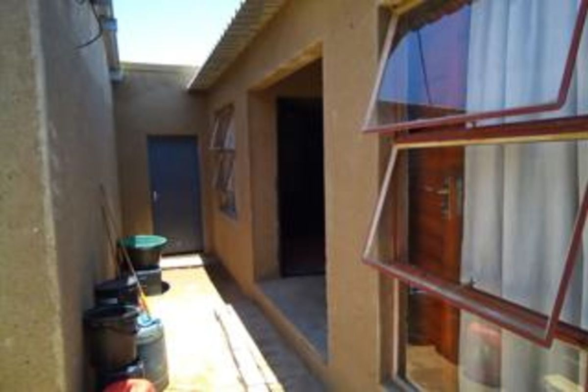 A narrow exterior side passage featuring plastered walls, top-hung windows, and a rear access door, showing signs of weathering and basic construction.