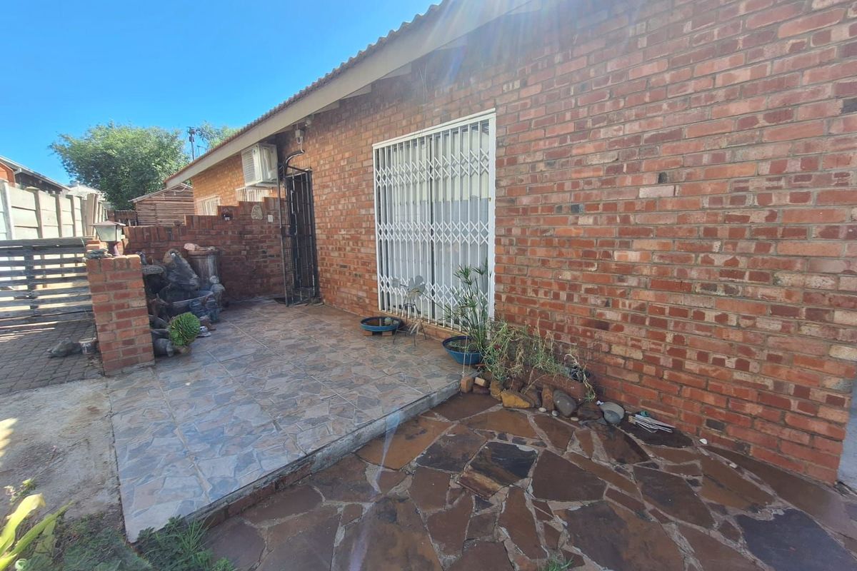 A rear exterior view of the house featuring a paved patio area, brick facade, and security gate access.