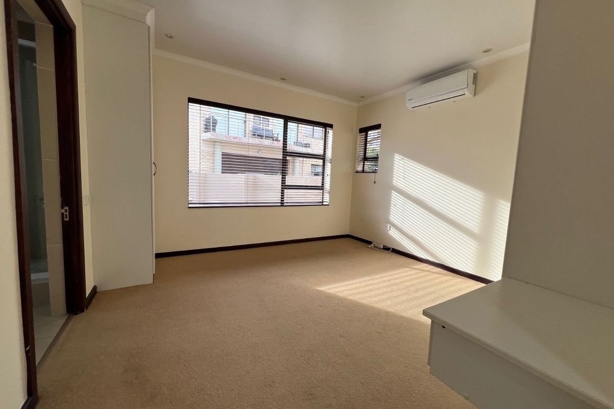 A bright, empty bedroom featuring neutral carpeting, built-in cupboards, a wall-mounted air conditioning unit, and large windows with fitted blinds.
