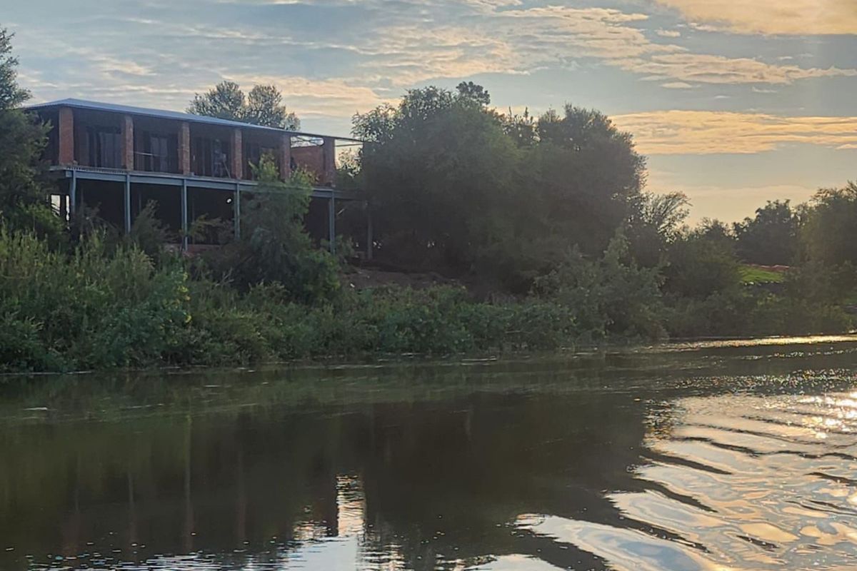 A view of an unfinished multi-level farm property situated along a riverbank, showcasing exposed brickwork and structural steel framing.