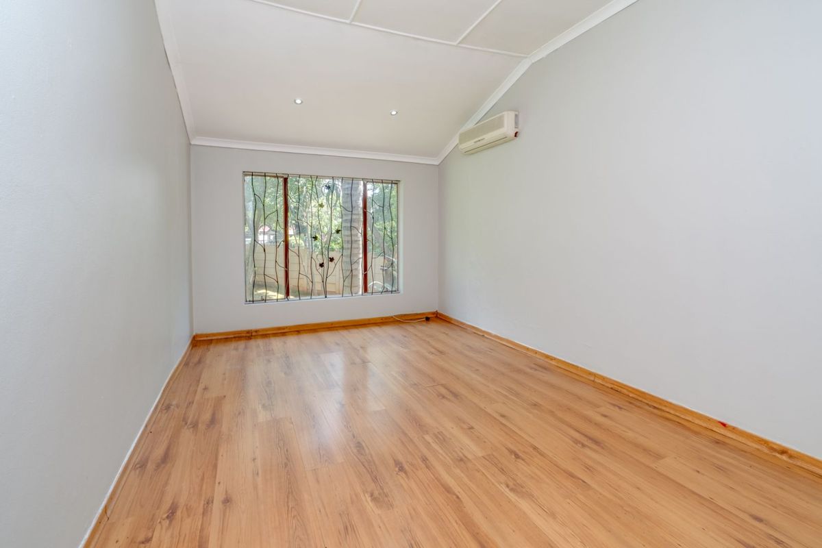 A bright, empty bedroom featuring light wood-look laminate flooring, neutral walls, a split-system air conditioning unit, and a window with decorative security bars.