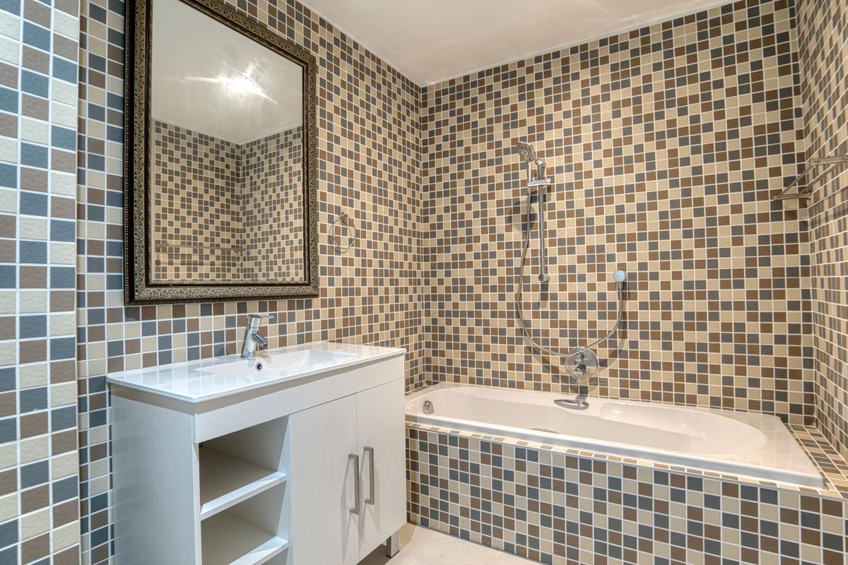 A functional bathroom featuring a built-in bathtub with shower mixer, a modern vanity unit with integrated basin, and decorative mosaic wall tiling.
