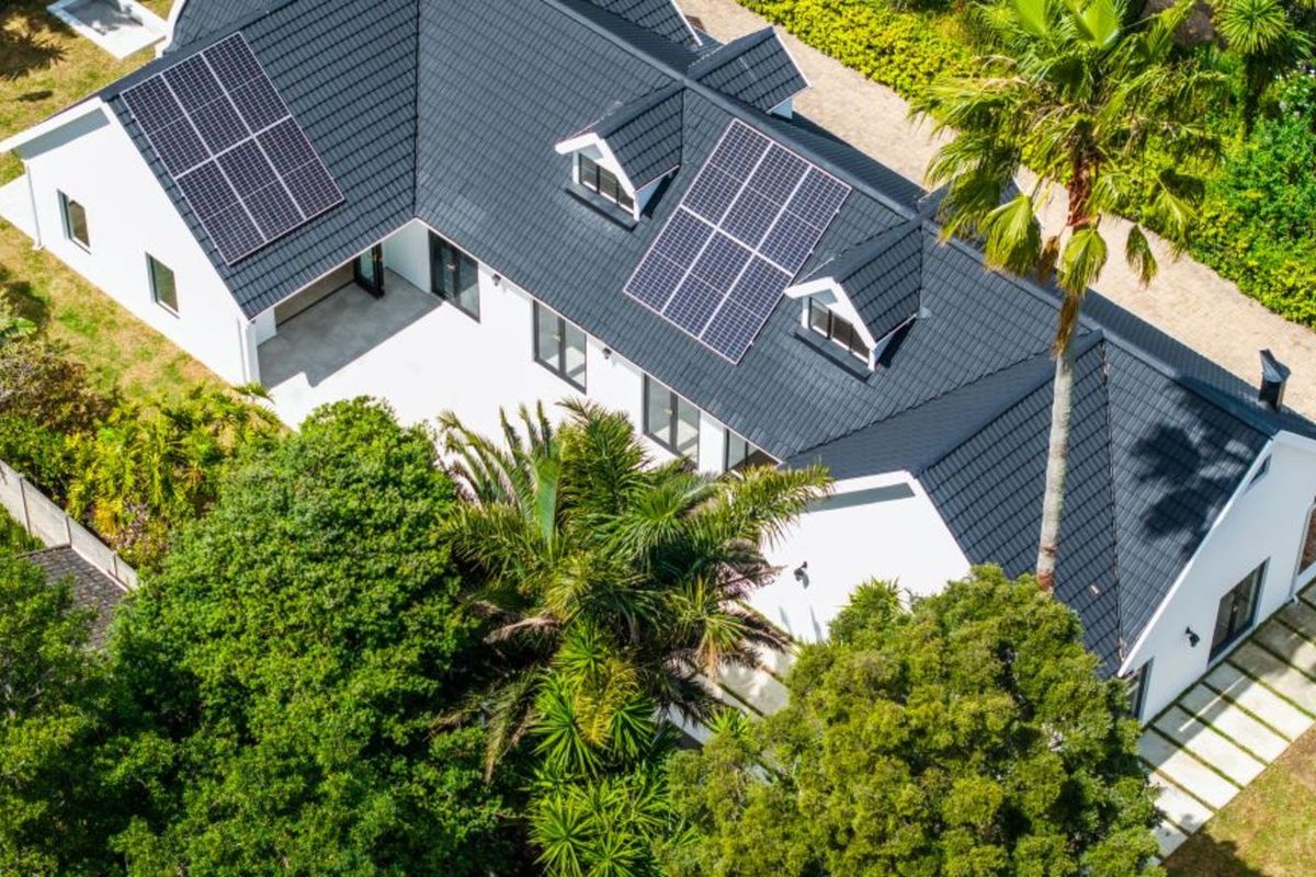 Aerial view of a modern house featuring a dark tiled roof with integrated solar panels, surrounded by lush greenery and landscaped garden areas.
