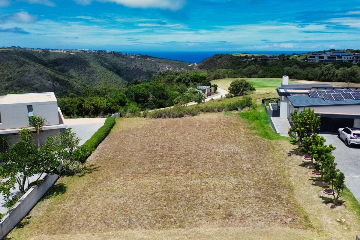 An elevated aerial view of a vacant residential plot situated between existing modern homes, featuring scenic landscape and distant ocean views.