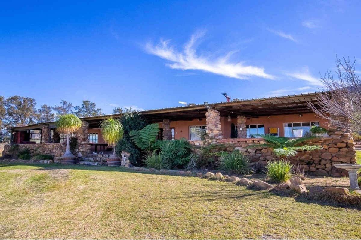 A rustic farm-style residence featuring extensive natural stone cladding, a covered veranda, and expansive surrounding lawns under a clear blue sky.