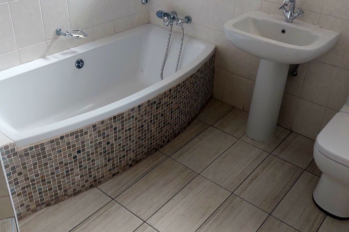 A functional bathroom featuring a tiled bathtub with mosaic detailing, a pedestal basin, and a standard toilet with neutral floor and wall tiling.