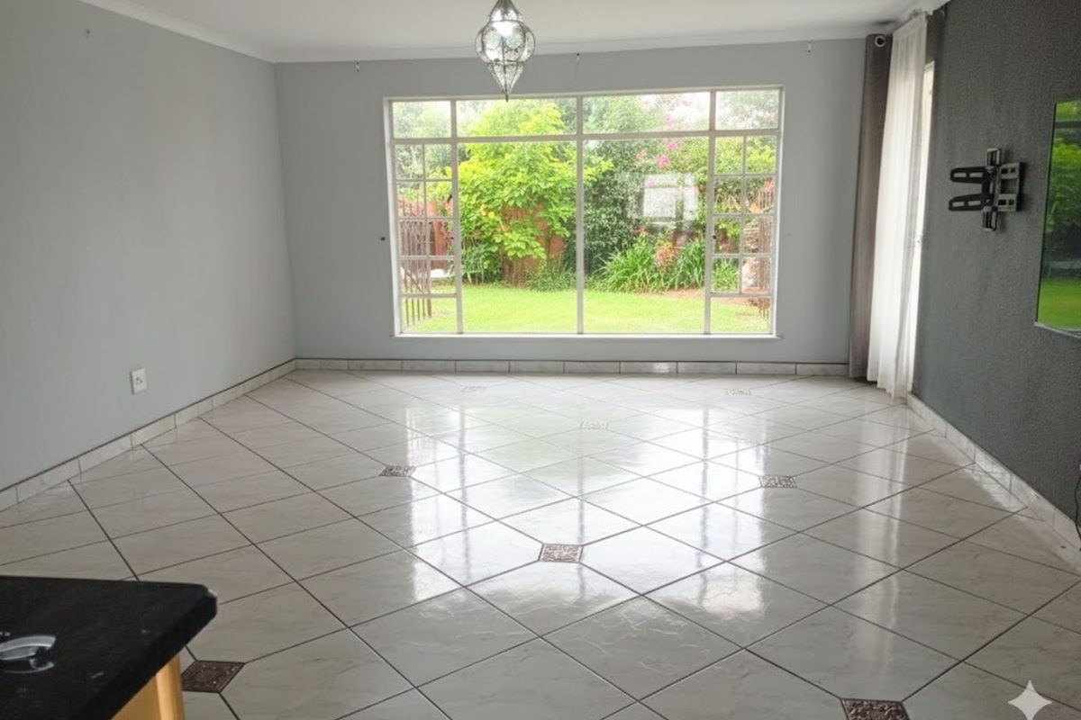 A spacious, empty living area featuring large windows with garden views, tiled flooring with decorative inlays, and a neutral color palette.