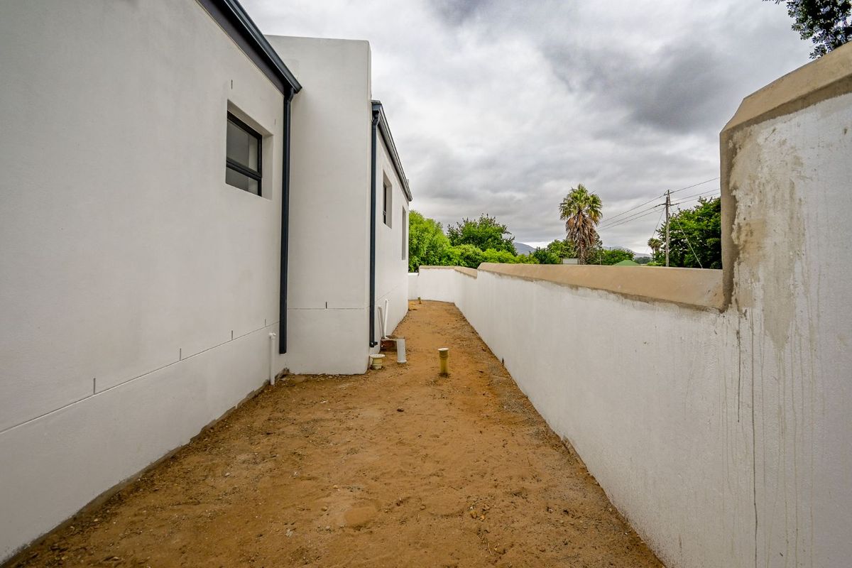 A narrow side exterior area featuring white rendered walls, exposed plumbing infrastructure, and an unfinished dirt ground surface.