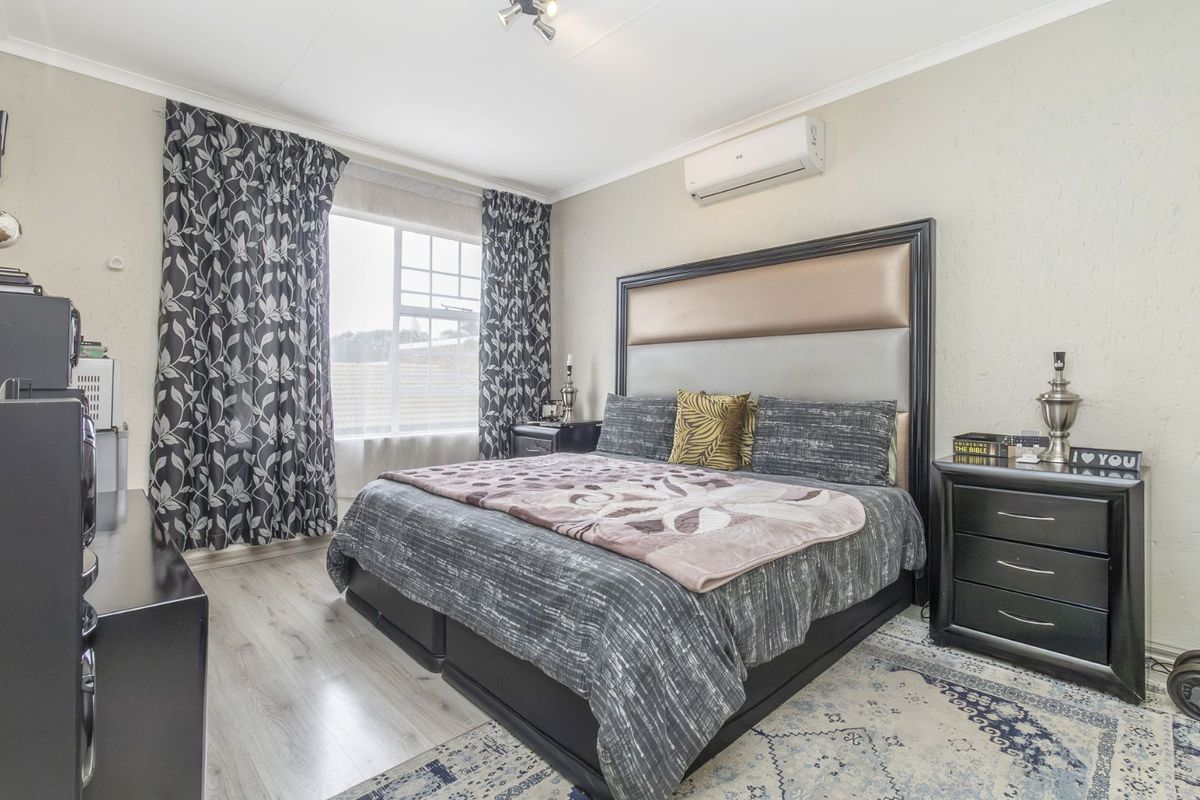 A well-lit bedroom featuring light-toned laminate flooring, a wall-mounted air conditioning unit, and a large window with curtain tracks.