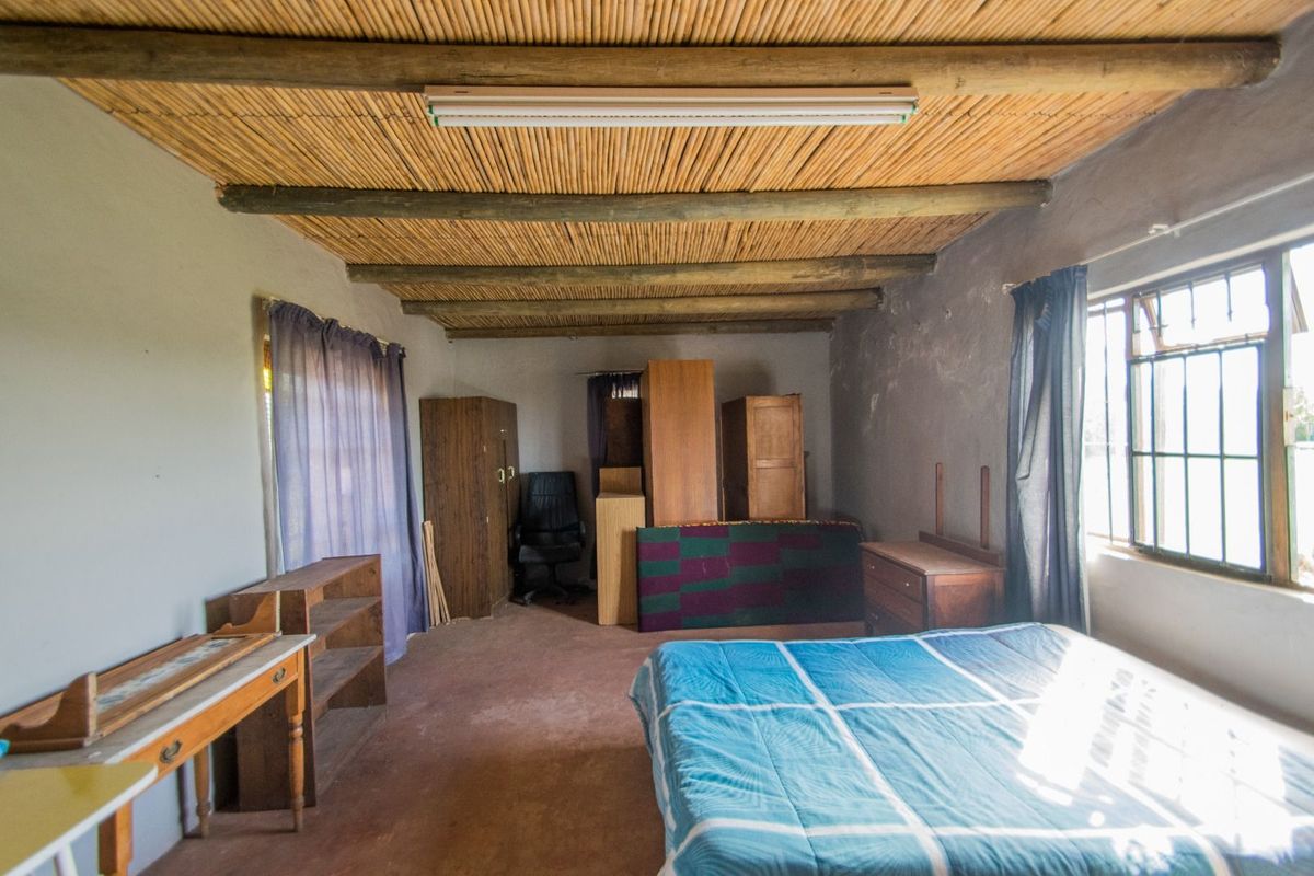 A rustic bedroom featuring a traditional reed and timber beam ceiling, large windows, and basic wall finishes.