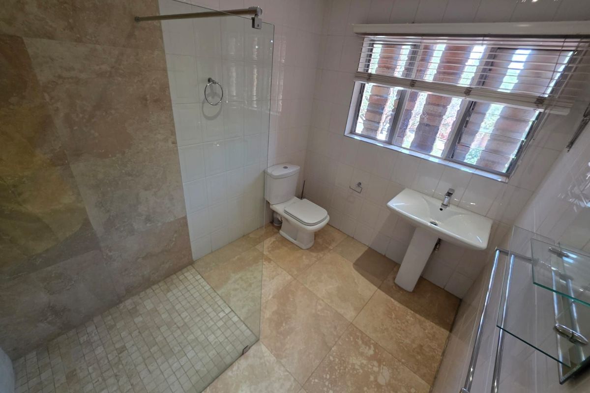A functional bathroom featuring a walk-in glass shower, pedestal basin, and toilet with neutral floor and wall tiling.
