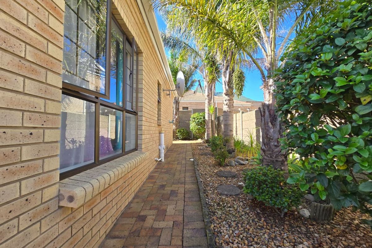 A side exterior view of the property featuring a paved walkway, brick facade, and established garden landscaping with palm trees.