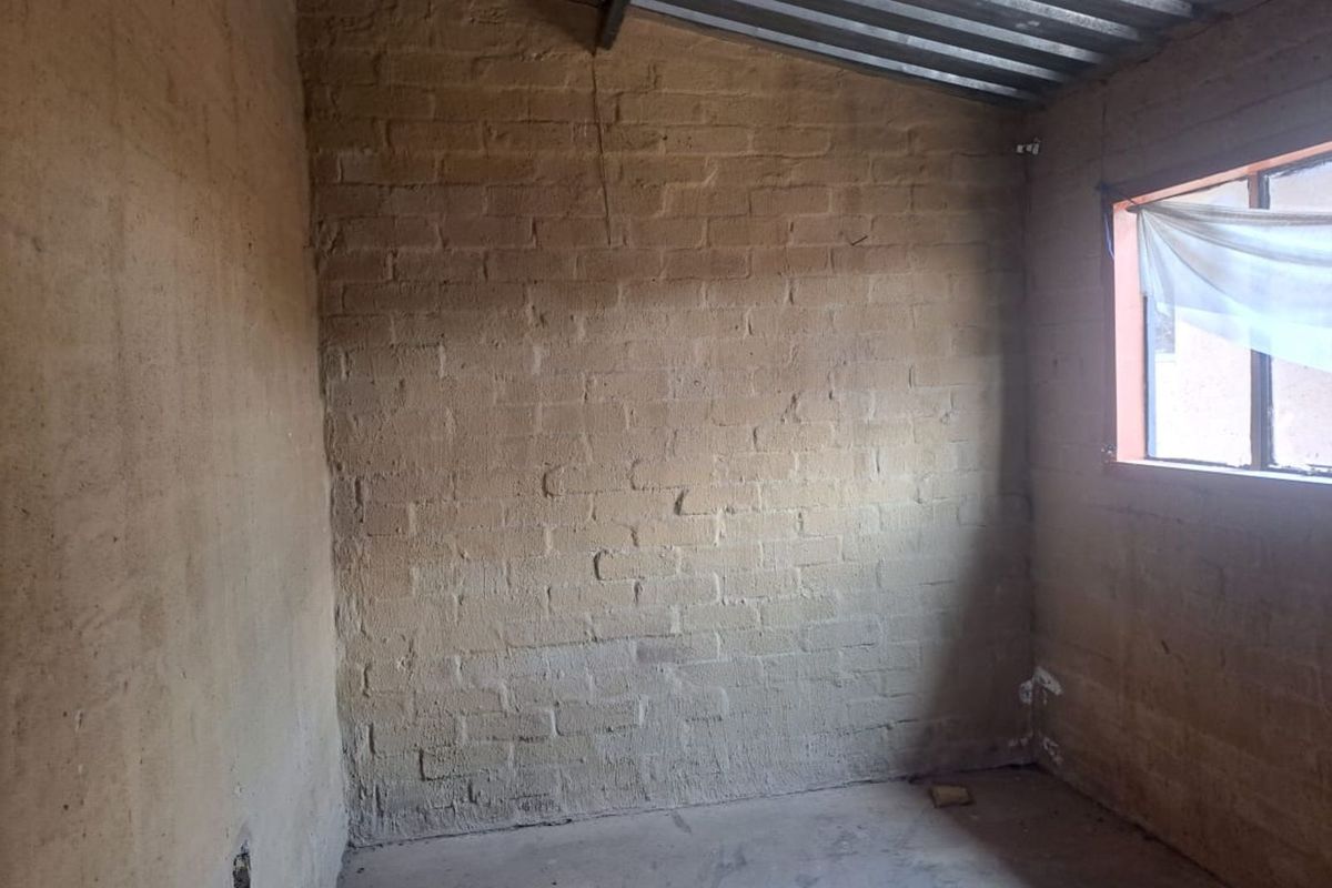 An unfinished interior room featuring exposed brick walls, a concrete floor, and an exposed metal roof structure with a single window.
