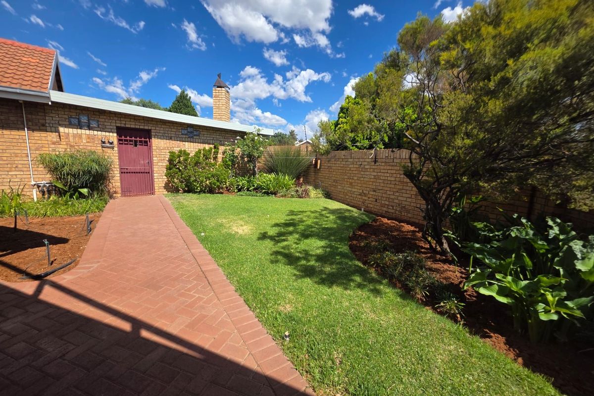 A well-maintained rear garden featuring a brick-paved pathway, established lawn, perimeter brick walling, and a brick chimney stack on the exterior facade.