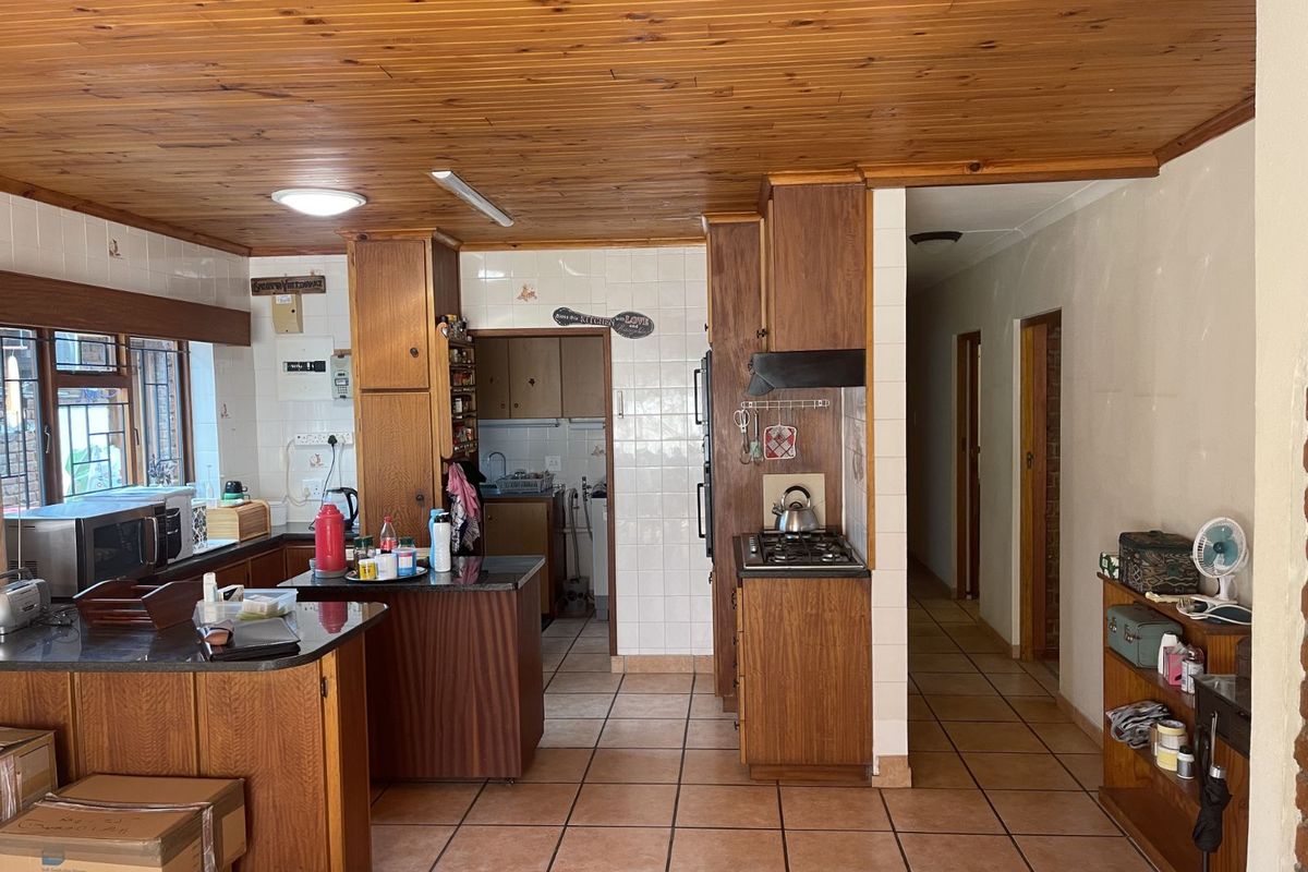 A functional kitchen featuring wood cabinetry, tiled splashbacks, and a connected scullery area, leading into a hallway with tiled flooring throughout.