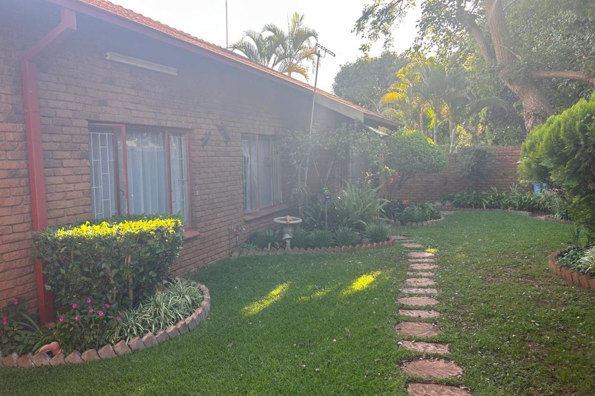 A view of the rear garden featuring a brick exterior wall, established landscaping, and a stone pathway leading through the lawn.