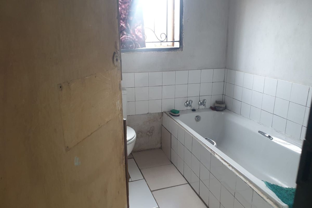 A dated bathroom featuring a built-in bathtub, tiled splashbacks, and a standard toilet, requiring cosmetic updates and repairs to wall finishes.