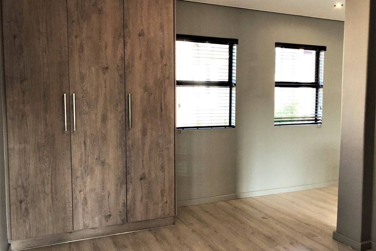A bright bedroom featuring modern wood-look laminate flooring, built-in wooden cabinetry, and dual windows with fitted blinds.