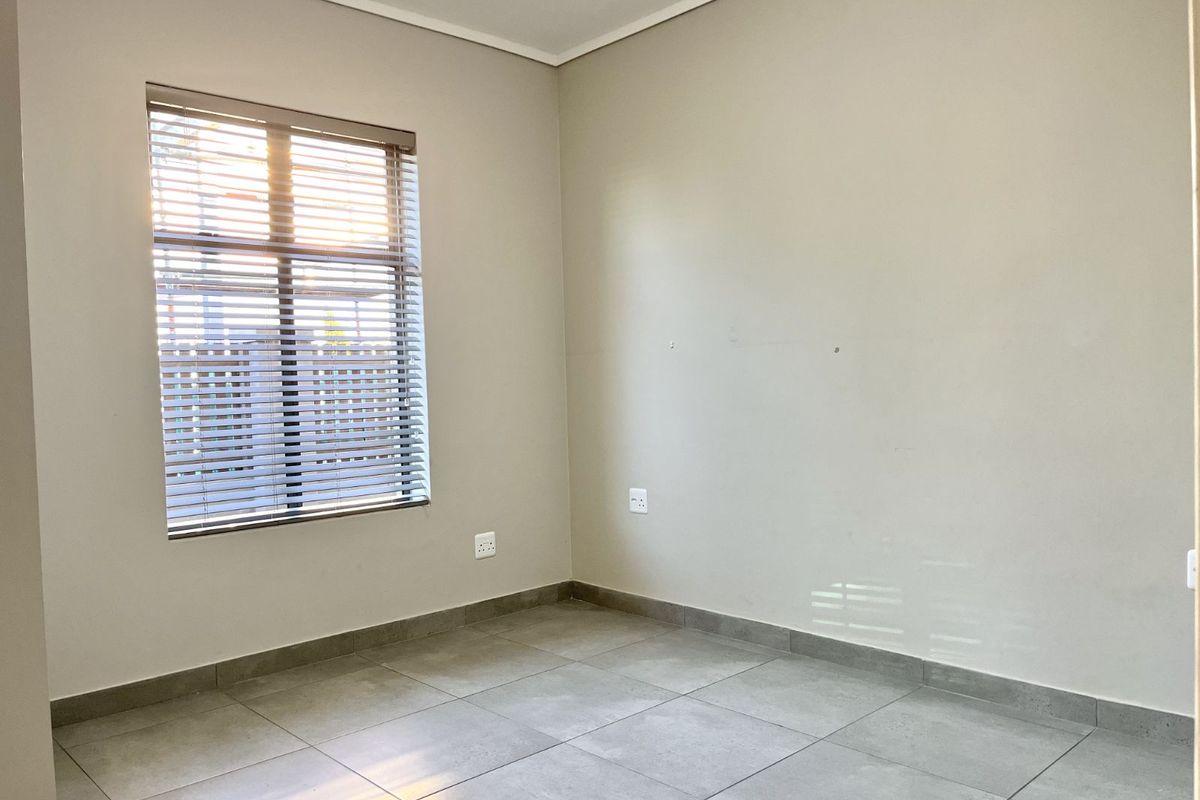 A bright, empty bedroom featuring neutral wall tones, large format floor tiles, and a window fitted with horizontal blinds.