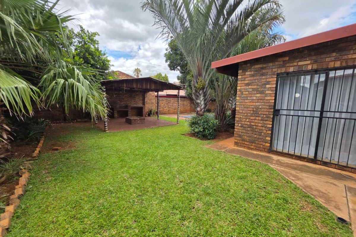 A private rear garden featuring a lawn, established palm trees, and a covered brick-built braai area for outdoor entertaining.